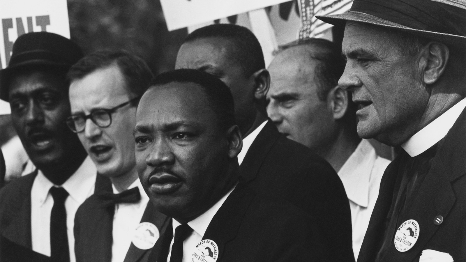 Black-and-white photograph of Martin Luther King Jr. at the 1963 March on Washington, standing among other civil rights leaders in front of protest placards, one reading "NOW!", with "March on Washington for Jobs & Freedom" buttons on their lapels