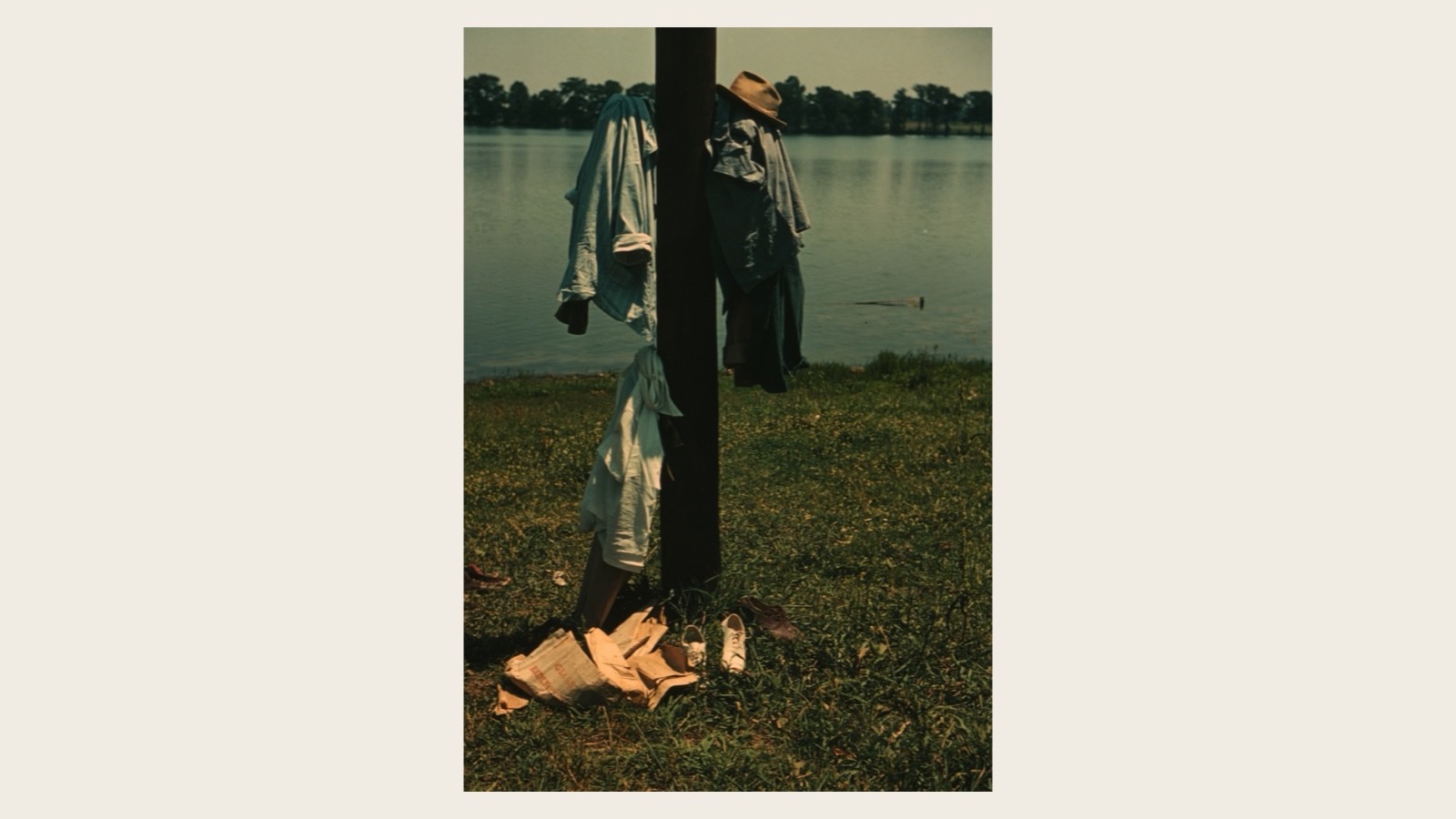 A 1940 color photograph of children's swimming clothes — pale shirts, a straw hat, and canvas sneakers — hung on a wooden telegraph pole beside a calm lake, the children themselves out of frame, off swimming in the water