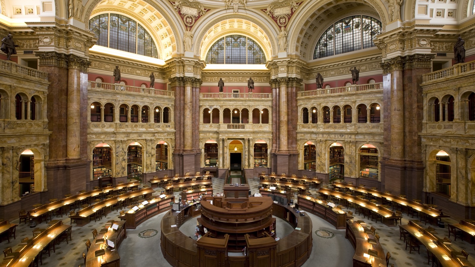 The Main Reading Room of the Library of Congress in Washington, D.C., a vast domed circular hall lined with tiers of books, with readers seated at wooden desks radiating from the center beneath ornate arches and columns