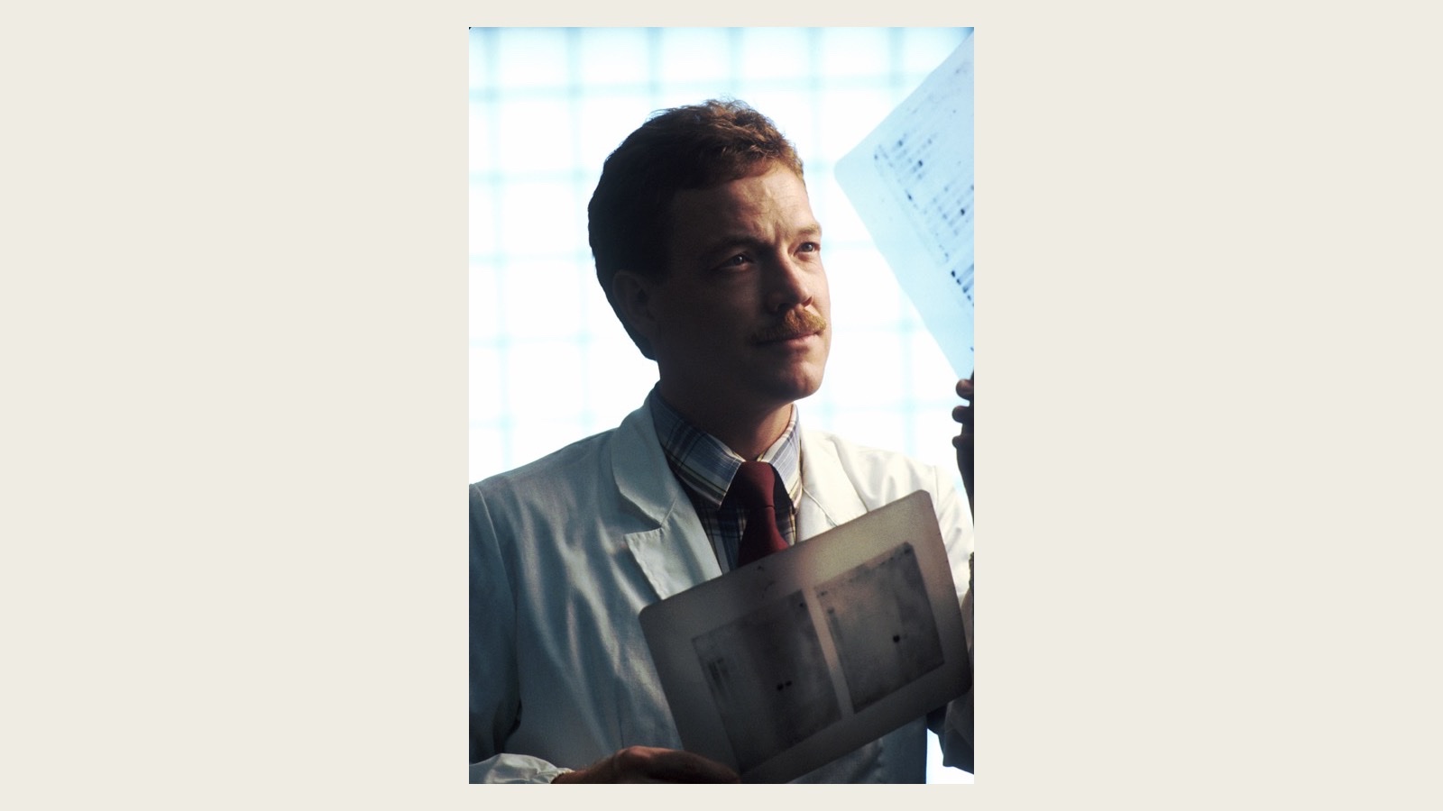 A photograph of a scientist in a white coat in 1989, holding a DNA sequencing autoradiograph film up to the light and studying it, with a second sequencing film in his other hand
