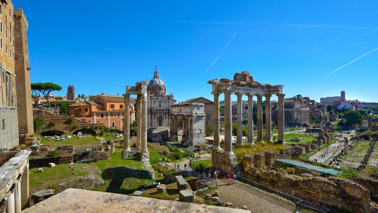 A photograph of the Roman Forum today: rows of weathered columns, including the tall columns of the Temple of Saturn and the Arch of Septimius Severus, standing among grassy ruins in the centre of Rome, with the Colosseum visible in the distance