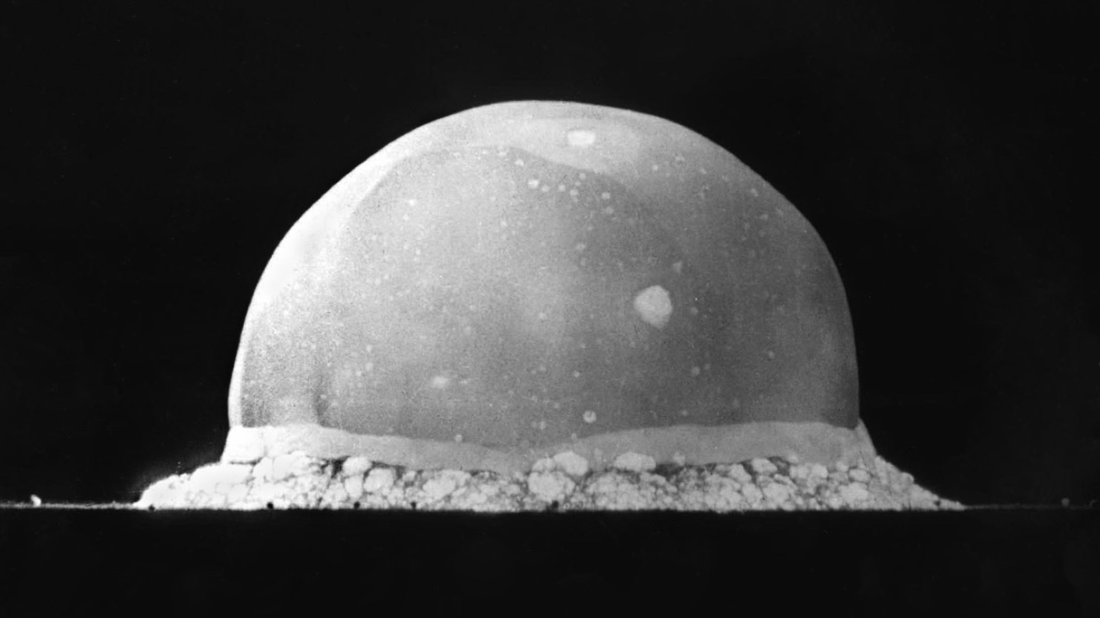 A black-and-white photograph of the Trinity nuclear test: a glowing, near-hemispherical dome of light rising from the desert floor in the first instant after the first atomic detonation in 1945